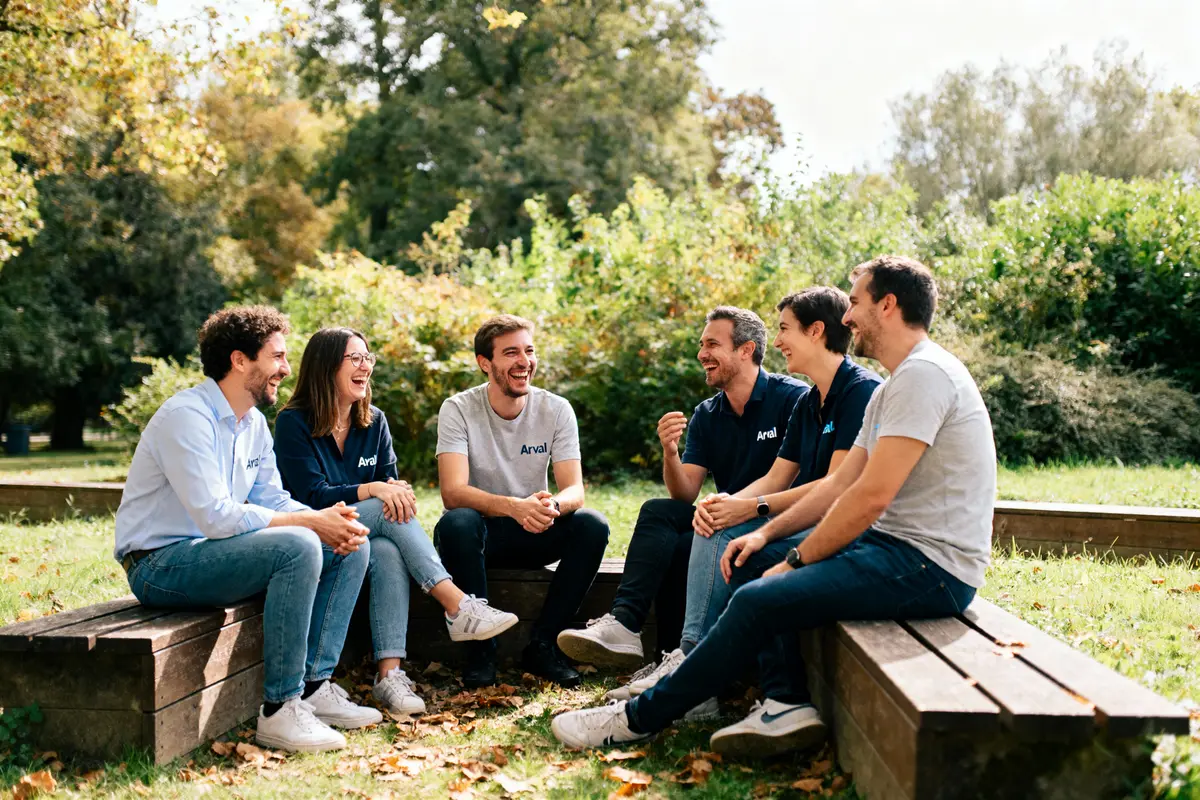 Un groupe de six personnes riant ensemble, assises sur des bancs en extérieur, entourées de verdure.