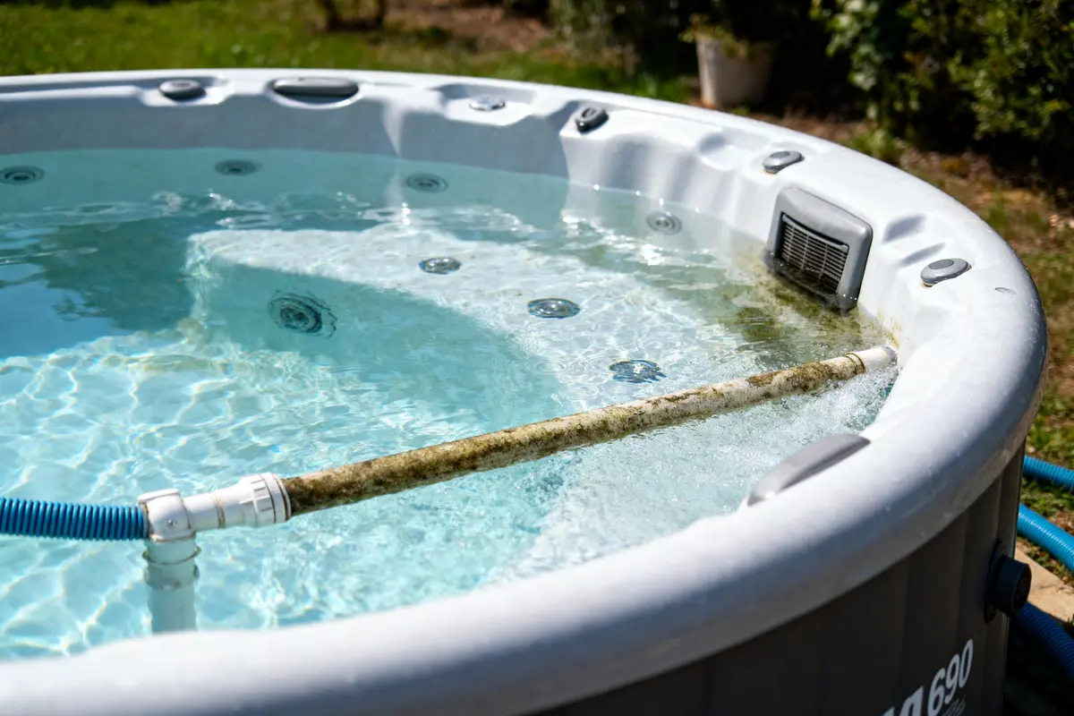 Jacuzzi extérieur ensoleillé avec tuyau d'aspiration et mousse, entouré de verdure et de pots.