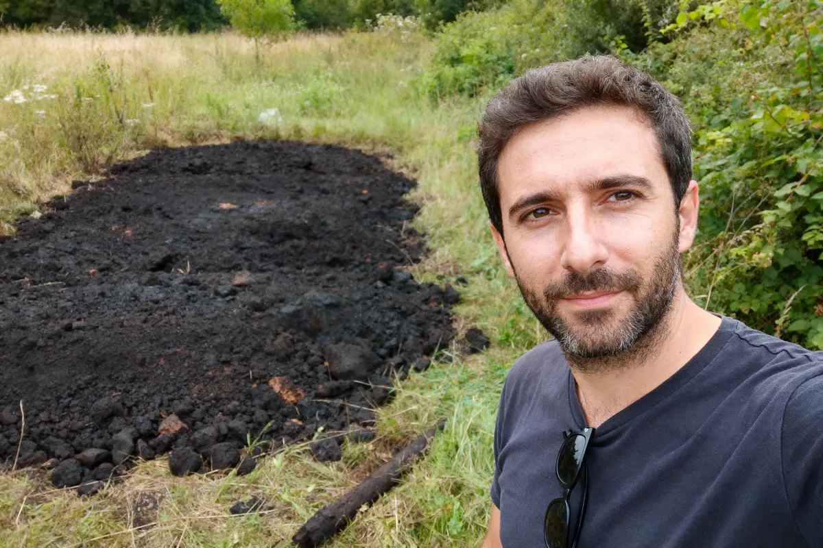Homme souriant en extérieur près d'un sol noirci, entouré de végétation verte.