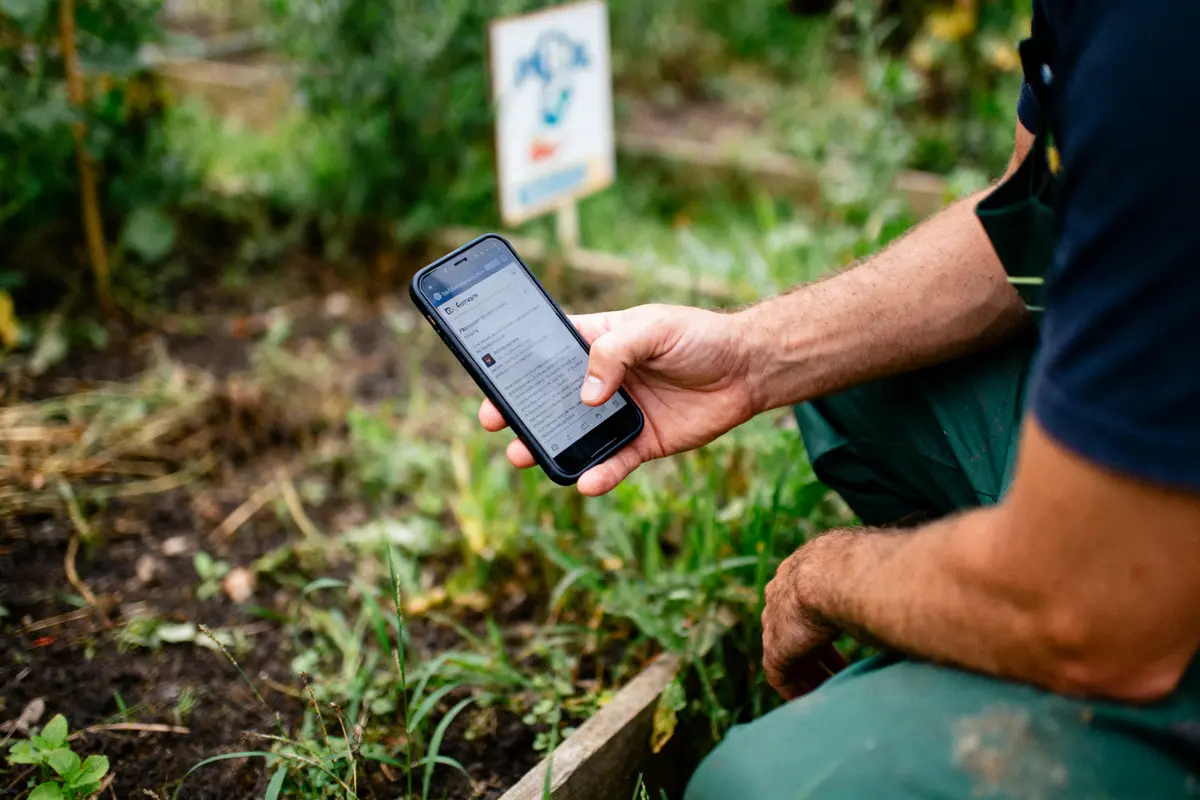 Personne utilisant un smartphone dans un jardin, avec des plantes et un panneau flou à l'arrière-plan.