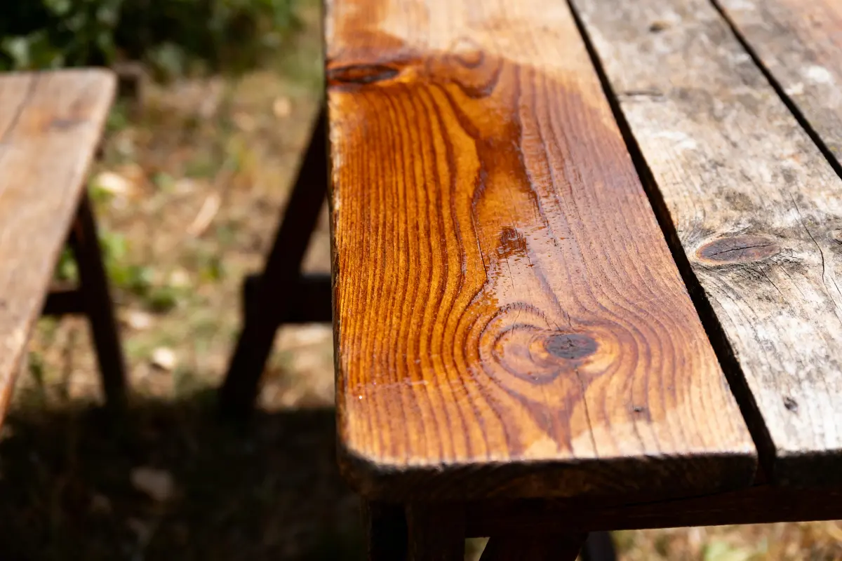 Table en bois partiellement mouillée, en extérieur, montrant texture et veinures distinctes sous la lumière du soleil.