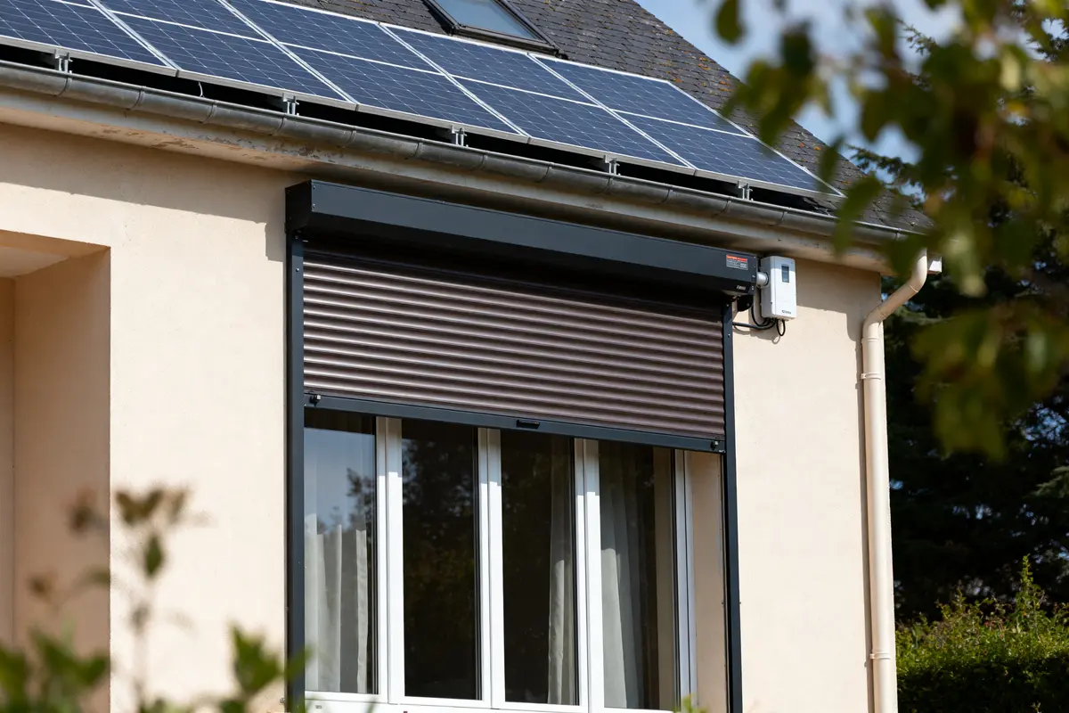 Maison avec volets roulants motorisés, panneaux solaires sur le toit, arbres au premier plan.