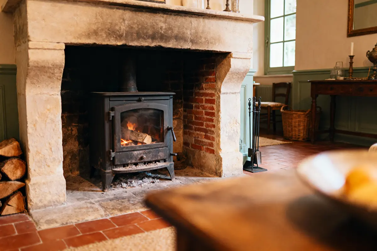 Poêle à bois allumé dans une cheminée en pierre et brique, pièce rustique avec parquet.