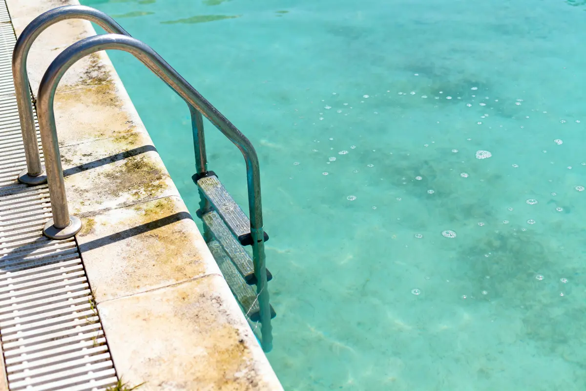 Échelle métallique de piscine, eau claire turquoise, margelles en pierre légèrement tachées et mousse.