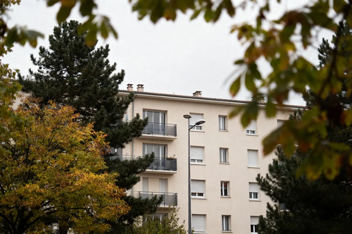Immeuble beige entouré d'arbres aux feuillages automnaux, lampadaire au premier plan, ciel couvert.