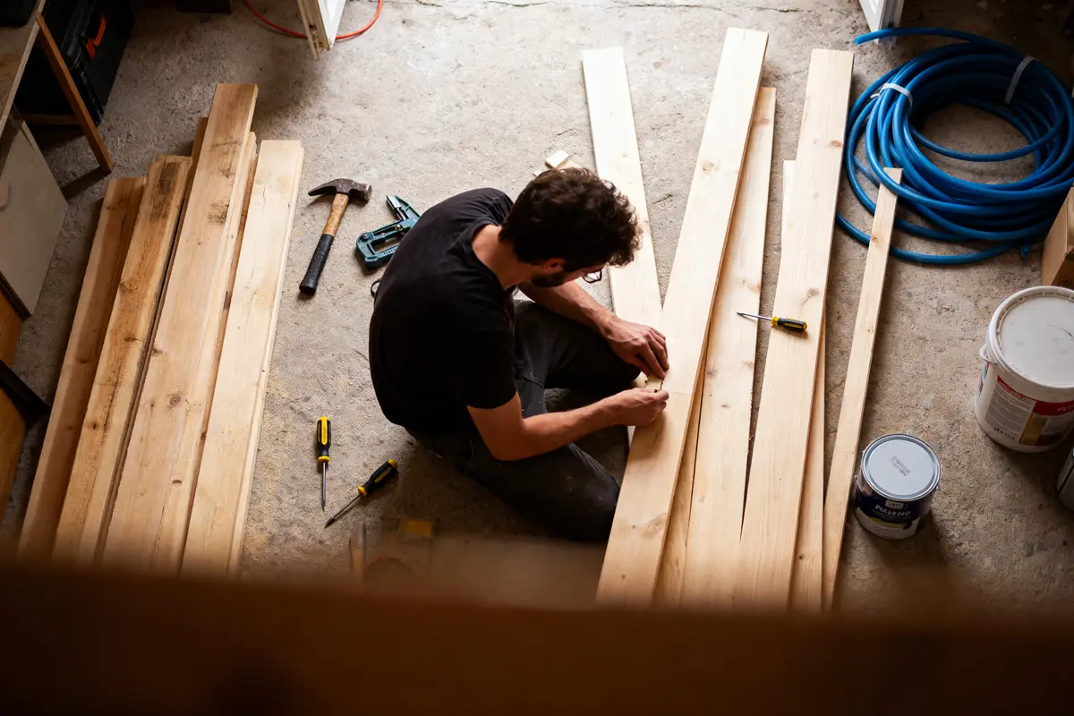 pexels-michal-pech-213601-757318 (1) Un homme travaille le bois, entouré d'outils, planches, marteau, tournevis dans un atelier.